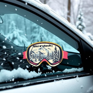 May include: A ski goggles sticker with the text "Ski o'clock!" features a mountain scene with pine trees. The sticker has a yellow, pink, and black colour scheme and is affixed to a car window with snow.