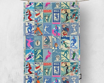 May include: A rectangular, vintage-style tea towel featuring a collage of retro ski posters in shades of blue, red, and pink. The posters depict skiers in action, with text in various languages. The towel is hanging from a wooden clothespin.
