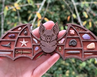 Puede incluir: Un expositor de madera con forma de murciélago con compartimentos para objetos pequeños. El expositor está pintado de marrón y presenta un murciélago tallado en el centro. Los compartimentos están llenos de varios objetos pequeños, como cristales, fósiles y conchas.