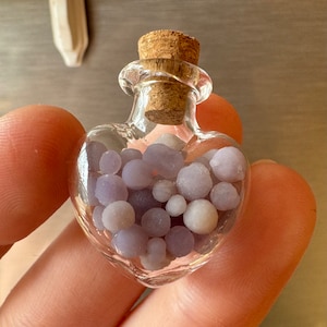 May include: A heart-shaped glass bottle filled with small, round, lavender and white beads. The bottle has a cork stopper. The clear glass allows the beads to be seen. The bottle is held in a person's hand.