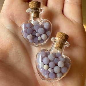 May include: Two heart-shaped glass bottles filled with small, round, lavender-colored beads. Each bottle has a cork stopper. The bottles are held in a hand.