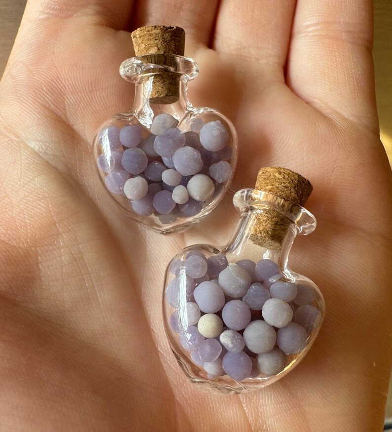 May include: Two heart-shaped glass bottles filled with small, round, lavender-colored beads. Each bottle has a cork stopper. The bottles are held in a hand.