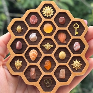 May include: A wooden honeycomb-shaped display tray with dark brown interior cells, each containing a small object including a gold bee, a gold key, a gold sun, a gold strawberry, a gold ladybug, various polished stones, and small seashells.