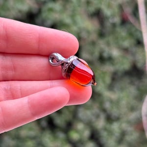 Könnte beinhalten: Ein handgefertigter Eichelanhänger mit silberfarbener Öse. Der Körper der Eichel hat eine transparente, leuchtend rot-orange Farbe mit einer dunkelbraunen Kappe. Der Anhänger wird in einer Hand vor einem verschwommenen grünen Hintergrund gehalten.