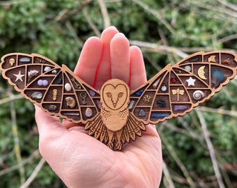 Puede incluir: Una escultura de búho de madera con detalles intrincados, que presenta un diseño de búho de granero con las alas extendidas. El cuerpo del búho está lleno de pequeños compartimentos, cada uno con un elemento natural diferente, como cristales, conchas y plumas.