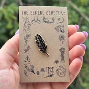 May include: A black and gold feather enamel pin attached to a rectangular kraft paper display card. The card features the text THE SERENE CEMETERY and TMS, surrounded by black line art illustrations of flowers, a spider, a skull, and a moth.