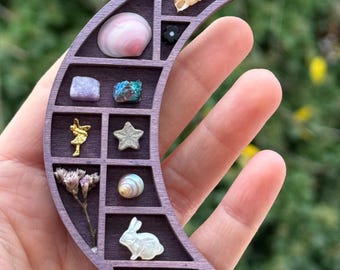 Puede incluir: Una caja de madera en forma de media luna con 12 compartimentos, cada uno lleno de un objeto pequeño, como una concha, un cristal, una flor y una pequeña figura de conejo.