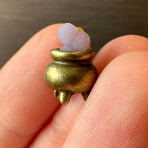 Könnte beinhalten: Ein kleiner goldener Kessel-Anhänger mit zwei violetten Zaubertrankflaschen darin. Der Anhänger wird in einer Hand gehalten.