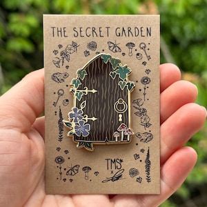 May include: Gold enamel pin depicting a wooden door with ivy, flowers, and mushrooms. The pin is displayed on a brown card with the text "The Secret Garden" and "TMS*" in black.