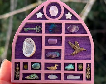 Puede incluir: Un expositor de madera rosa y morado con varios objetos pequeños. Presenta una llave, una pluma, cristales, un colibrí y otros elementos decorativos. El expositor se sostiene en una mano.