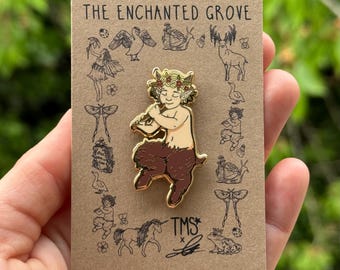 Può includere: Spilla in smalto dorato con una piccola figura maschile dalla pelle scura, con una corona di fiori, che suona un flauto. La figura ha gambe di capra ed è posizionata su uno sfondo marrone con illustrazioni di varie creature del bosco. Il testo "THE ENCHANTED GROVE" è stampato nella parte superiore della carta. Il testo "TMS*" è stampato nella parte inferiore della carta.