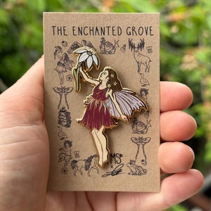 May include: Gold enamel pin featuring a fairy holding a white flower. The fairy has brown hair, a burgundy dress, and translucent wings. The pin is on a brown card with the text "The Enchanted Grove" and other illustrations.