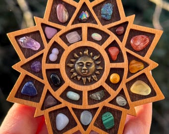 Puede incluir: Expositor de madera en forma de estrella con un centro con forma de sol, que contiene varias piedras preciosas de colores. El expositor es de madera marrón claro y se sostiene en una mano sobre un fondo verde borroso. Las piedras preciosas son de diferentes formas y tamaños.