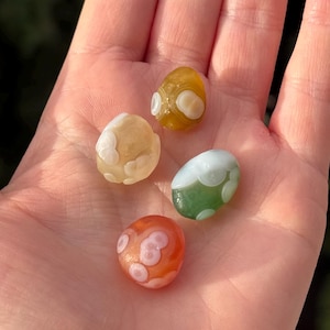May include: A hand holds four polished gemstone beads. The beads are oval-shaped and feature a variety of colours, including orange, yellow, and green, with white, cloud-like patterns. These beads are likely used for jewellery making or as decorative elements.