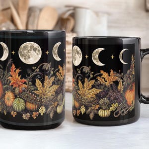 May include: Two black ceramic mugs with a moon and autumn leaves design. The mugs feature a full moon, crescent moons, and stars against a black background. The design also includes autumn leaves, vines, and pumpkins.