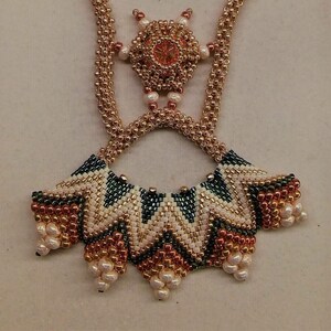 May include: A gold, white, and green beaded necklace with a geometric design. The necklace has a pendant with a starburst pattern.