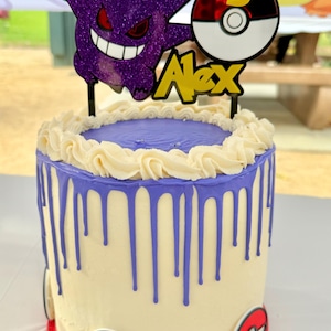 May include: A birthday cake featuring a purple Gengar cake topper, a Poke Ball with the number 5, and the name "Alex" in yellow. The cake has a white base with purple drip icing and Poke Ball decorations.