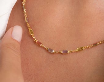 May include: A delicate gold necklace featuring small, rectangular gemstones in shades of pink, yellow, and purple. The necklace is worn around the neck, with a hand gently touching the chain. The gemstones are approximately 0.5 cm in length.