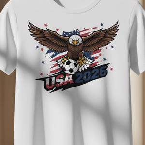 Puede incluir: Camiseta blanca con un diseño gráfico de un águila con las alas extendidas, un balón de fútbol y el texto "USA 2026". El diseño incorpora los colores de la bandera estadounidense. El águila es el foco central.