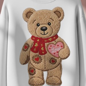 Peut inclure: Sweat-shirt blanc avec un motif d'ours en peluche. L'ours est brun, porte une écharpe rouge et tient un cœur rose avec le mot "Love". L'ours a des motifs de cœurs et de fleurs. Un design mignon et confortable.