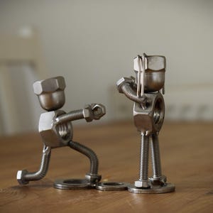 Op de afbeelding: Twee metalen sculpturen die figuren in een bokswedstrijd uitbeelden. De figuren zijn gemaakt van moeren, bouten en ringen, met de hoofden van grote zeskantmoeren. Geplaatst op een houten oppervlak.