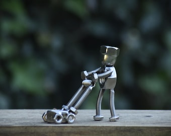 May include: A handcrafted metal sculpture of a person pushing a lawnmower. The figure is made from nuts and bolts, with a nut for the head and bolts for the arms, legs, and body. The lawnmower is also made of metal.