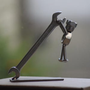 Puede incluir: Una escultura metálica plateada de una llave inglesa sosteniendo una figura hecha de tuercas y pernos. La llave inglesa se apoya sobre una pequeña base metálica. La figura parece estar sosteniendo una tuerca. El fondo está borroso.