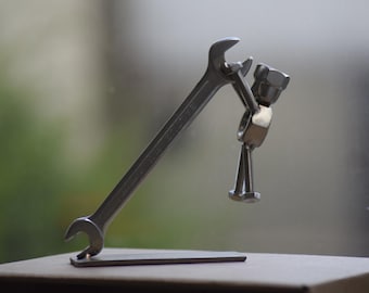 May include: A metal sculpture of a wrench holding a figure made of nuts and bolts. The wrench is silver and stands upright on a small metal base. The figure appears to be holding a nut. The background is blurred.