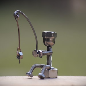 May include: A metal sculpture of a fisherman made from nuts, bolts, and wire. The figure is seated, holding a fishing rod with a hook. The sculpture is silver-colored and sits on a wooden surface against a blurred green background.