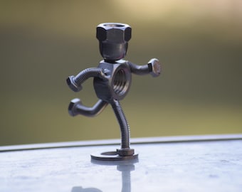 May include: A metal sculpture of a running figure, crafted from nuts, bolts, and a spring. The figure has a hexagonal nut head, a nut body, and bolts for arms and legs. The sculpture is on a round metal base.