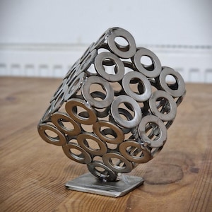 Könnte beinhalten: Eine geometrische Metallskulptur, die aus zahlreichen kreisförmigen Unterlegscheiben zusammengeschweißt wurde. Die Skulptur ist auf einem quadratischen Metallsockel montiert und steht auf einer Holzoberfläche. Das Metall hat eine silberne Farbe.