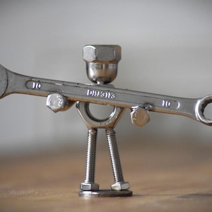 Puede incluir: Una escultura caprichosa hecha de hardware metálico, que se asemeja a una figura. Una llave inglesa forma el cuerpo, con pernos y tuercas utilizados para la cabeza, los brazos y las piernas. La llave inglesa está marcada con el número 10 y el texto "DIN3113".