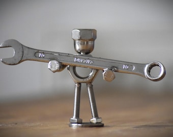 May include: A whimsical sculpture crafted from metal hardware, resembling a figure. A wrench forms the body, with bolts and nuts used for the head, arms, and legs. The wrench is marked with the number 10 and the text "DIN3113".