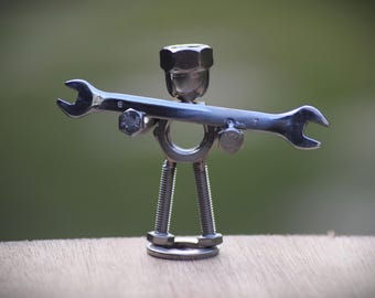May include: A metal sculpture of a figure made from hardware. The head is a large nut, the body is a ring, and the legs are threaded rods. A wrench is held across the shoulders. The sculpture is on a wooden surface.