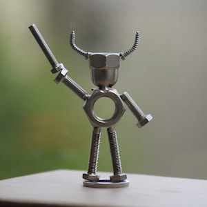 May include: A handcrafted metal sculpture of a figure made from nuts, bolts, and screws. The figure has a nut for a head, bolts for arms and legs, and a screw held as a weapon. The sculpture is silver and stands on a small metal base.