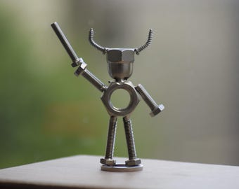 May include: A handcrafted metal sculpture of a figure made from nuts, bolts, and screws. The figure has a nut for a head, bolts for arms and legs, and a screw held as a weapon. The sculpture is silver and stands on a small metal base.