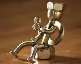 May include: A handcrafted metal sculpture of a person sitting, made from nuts, bolts, and washers. The figure is seated on a large nut, with bolt legs and arms, and a nut head. The sculpture is a unique piece of industrial art.