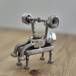 Puede incluir: Escultura metálica artesanal de un levantador de pesas. La figura está hecha de tuercas, pernos y tornillos, con una barra sobre la cabeza. La escultura está sentada en un banco de metal. Una obra de arte única de estilo industrial.