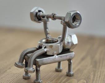 May include: A handcrafted metal sculpture of a weightlifter. The figure is made from nuts, bolts, and screws, with a barbell overhead. The sculpture is sitting on a bench made of metal. The piece is a unique, industrial-style art.