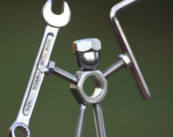 May include: A small figurine constructed from silver-toned metal hardware, including nuts, bolts, and washers. The figure features a nut for a head, a larger nut for a torso, and bolt legs. It holds a metal wrench in one hand and an L-shaped hex key in the other.