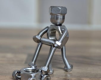 May include: A small figurine constructed from silver-colored metal hardware, including threaded bolts, nuts, and screws. The figure is shaped like a person sitting down and leaning forward, with its hands resting on a larger hexagonal nut base.