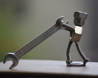May include: A whimsical sculpture of a figure constructed from metal hardware. The body is made of a nut, bolt, and washer, with a wrench held as if the figure is carrying it. The sculpture is silver and sits on a neutral surface.