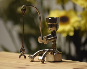 Puede incluir: Una pequeña figura construida con componentes de ferretería de metal plateado, incluyendo tuercas, pernos y tornillos. La figura tiene forma de persona sentada que sostiene una caña de pescar hecha de un alambre de metal fino con un pequeño anzuelo.