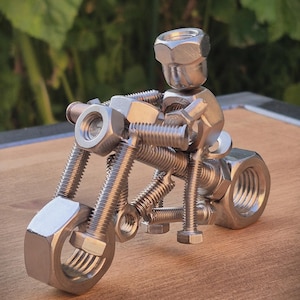 Può includere: Una piccola scultura di una motocicletta costruita con componenti metallici color argento, inclusi dadi esagonali, bulloni filettati e viti di varie dimensioni. La figura presenta un grande dado come ruota anteriore e posteriore.
