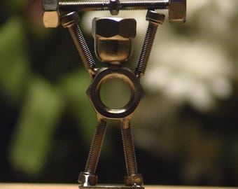 May include: A small figurine constructed from silver-coloured metal hardware, including nuts, bolts, and threaded rods. The figure is posed with arms raised, holding a barbell made of a threaded rod with a nut at each end above its head.