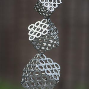Könnte beinhalten: Eine moderne Metallskulptur, die aus drei gestapelten, rautenförmigen Formen besteht. Jede Form besteht aus zahlreichen silbernen Unterlegscheiben, wodurch eine einzigartige, industrielle Ästhetik entsteht. Die Skulptur steht auf einem kleinen, kreisförmigen Sockel.