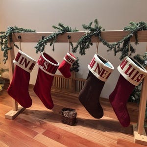 Può includere: Un portacalze in legno con cinque calze di Natale. Le calze sono rosse e marroni con polsini bianchi e nomi ricamati. Il supporto è decorato con ghirlanda sempreverde.