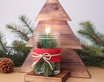 May include: A wooden Christmas tree candle holder with a glass jar filled with greenery and a red ribbon. The jar has a lit candle inside.