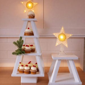 Könnte beinhalten: Ein weißer, mehrstufiger Cupcake-Ständer in Form eines Weihnachtsbaums mit Cupcakes, die mit weißem Frosting und roten Himbeeren verziert sind. Eine weiße sternförmige Leuchte sitzt oben auf dem Ständer. Eine zweite weiße sternförmige Leuchte sitzt auf einem weißen, mehrstufigen Ständer.
