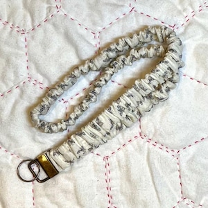 May include: A fabric lanyard featuring a gathered, scrunchie-style design in an off-white color with a repeating pattern of small, dark gray bees. The lanyard includes a metallic gold-toned rectangular clasp and a circular ring at the end.